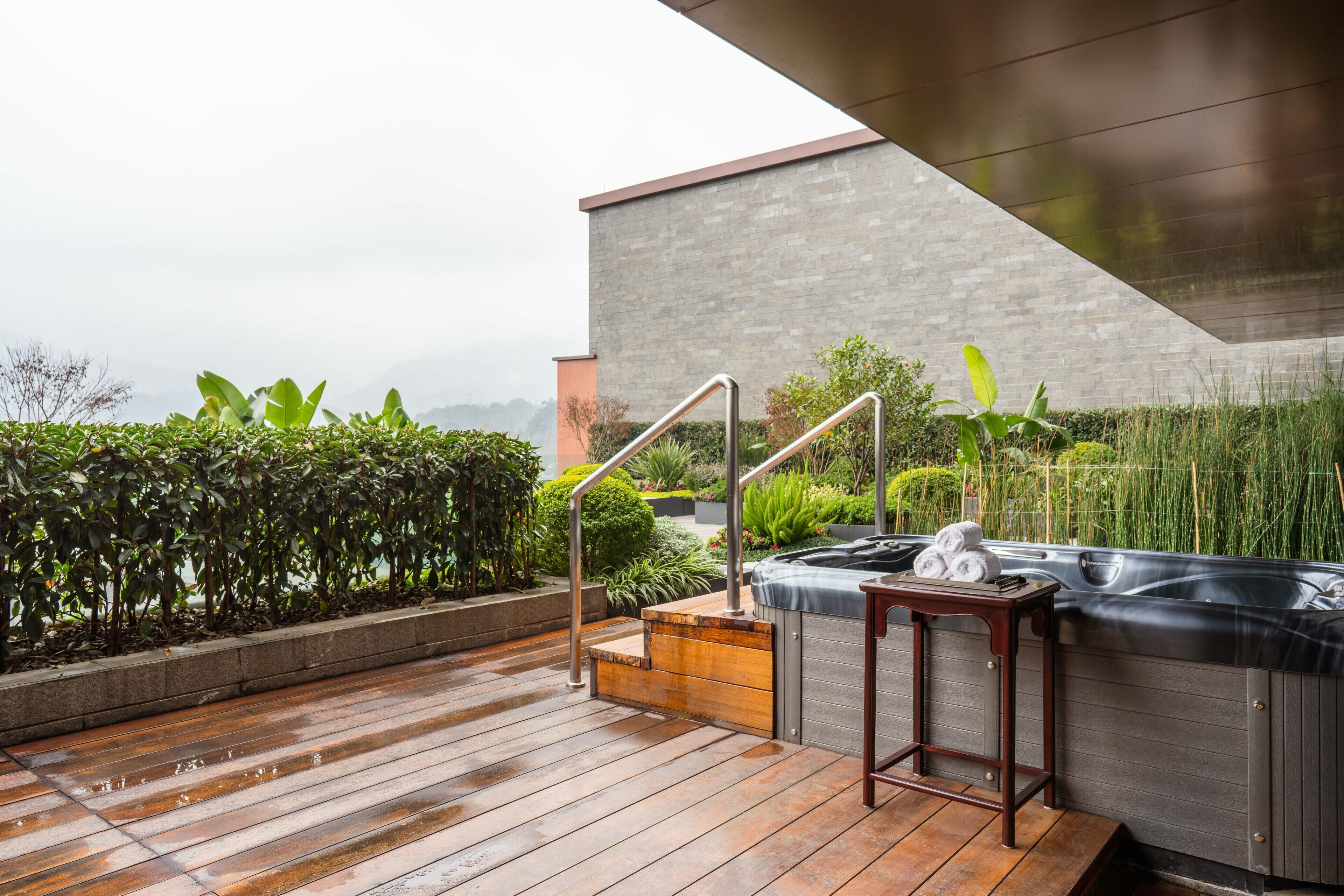 A wooden deck with a hot tub and a table, surrounded by greenery and a modern building.