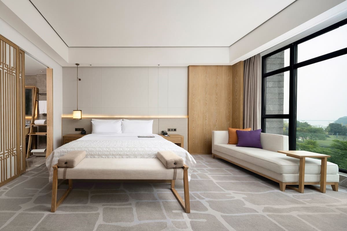A modern hotel room with a large bed, a sofa, a small table, and a view of the outdoors.