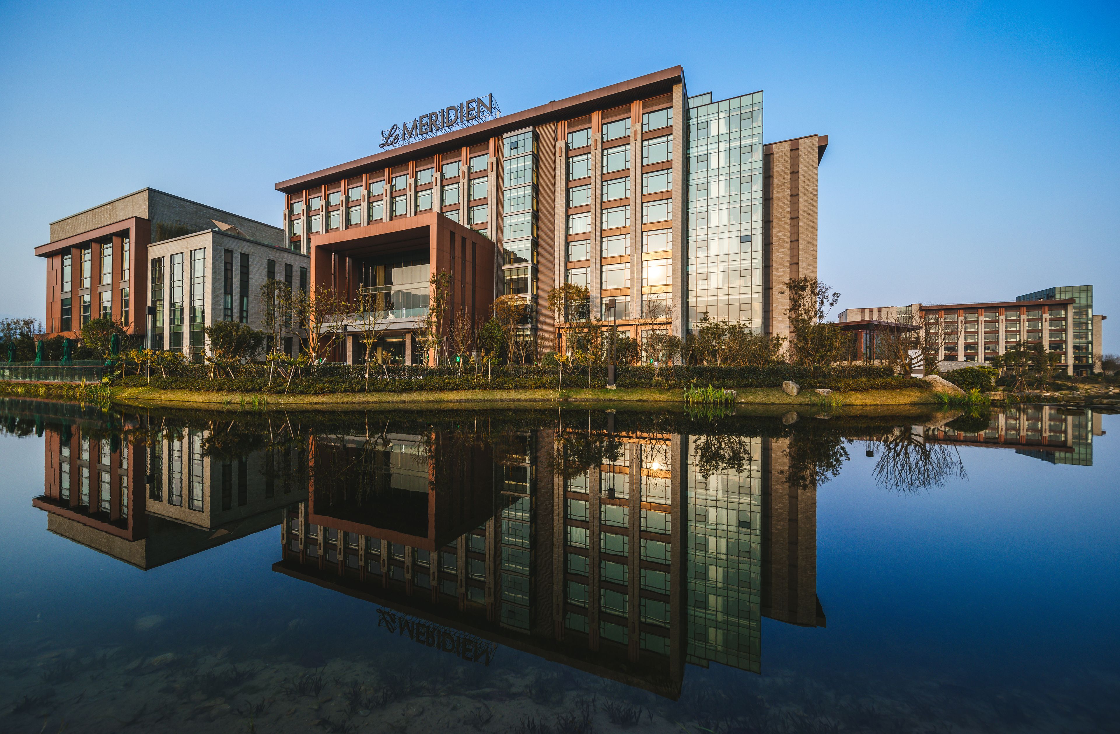 Un hôtel moderne à la façade vitrée, qui se reflète dans l’eau, et sur lequel on peut lire l’inscription « MERIDIEN ».