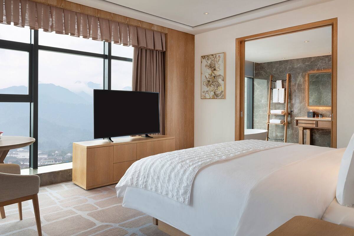 A hotel room with a large bed, a television on a wooden cabinet, a dining table, and a view of mountains through large windows.
