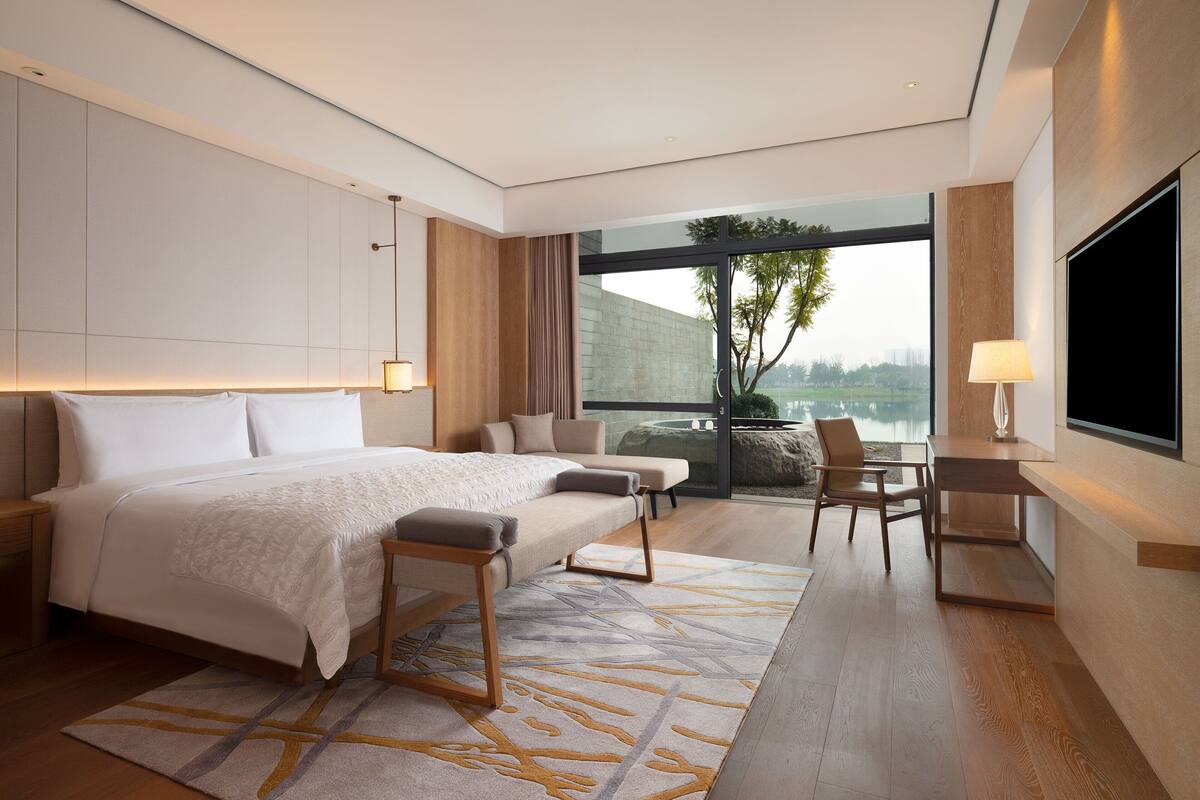 A modern hotel room with a large bed, a desk, and a view of a lake.