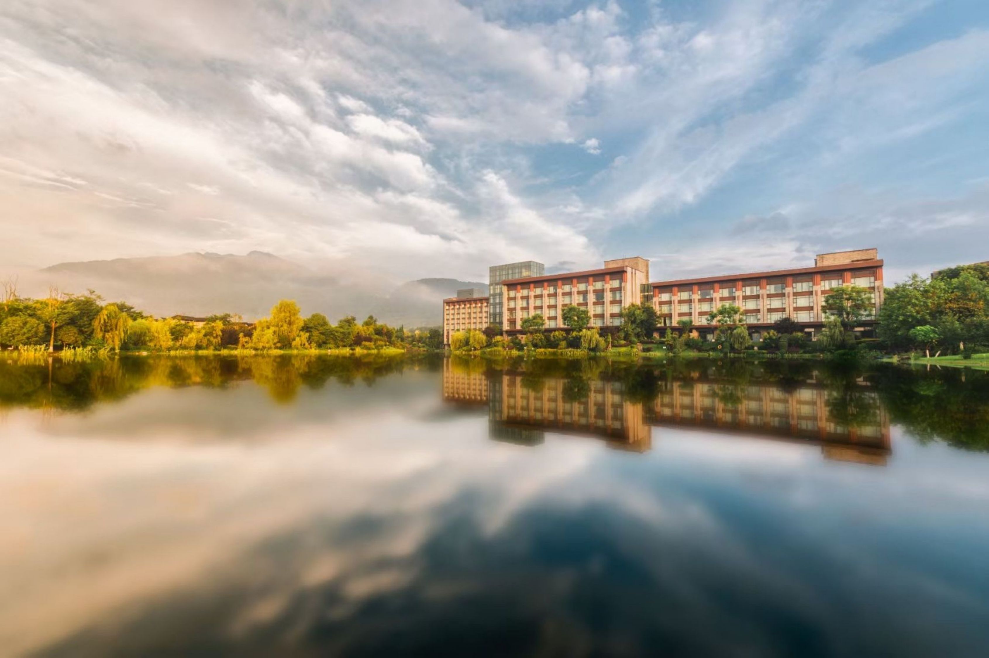 Un hotel moderno con su reflejo en un lago tranquilo, rodeado de vegetación bajo un cielo parcialmente nublado.