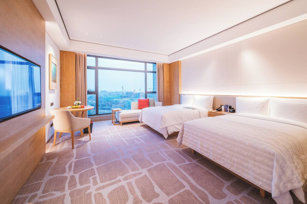 A hotel room with two beds, a desk, a chair, and a large window offering a city view.