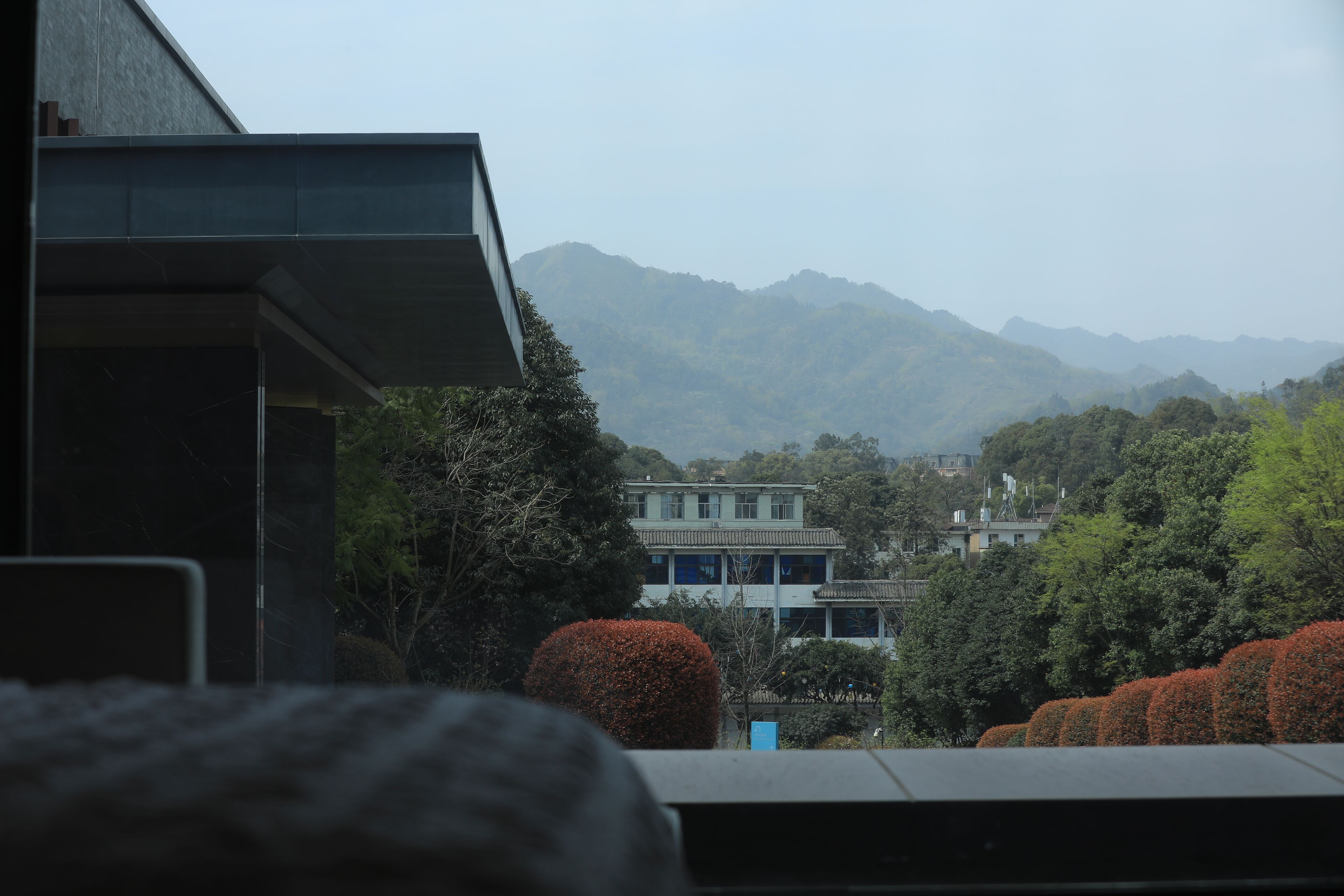 A modern building with a flat roof and large windows, surrounded by lush greenery and mountains in the background.