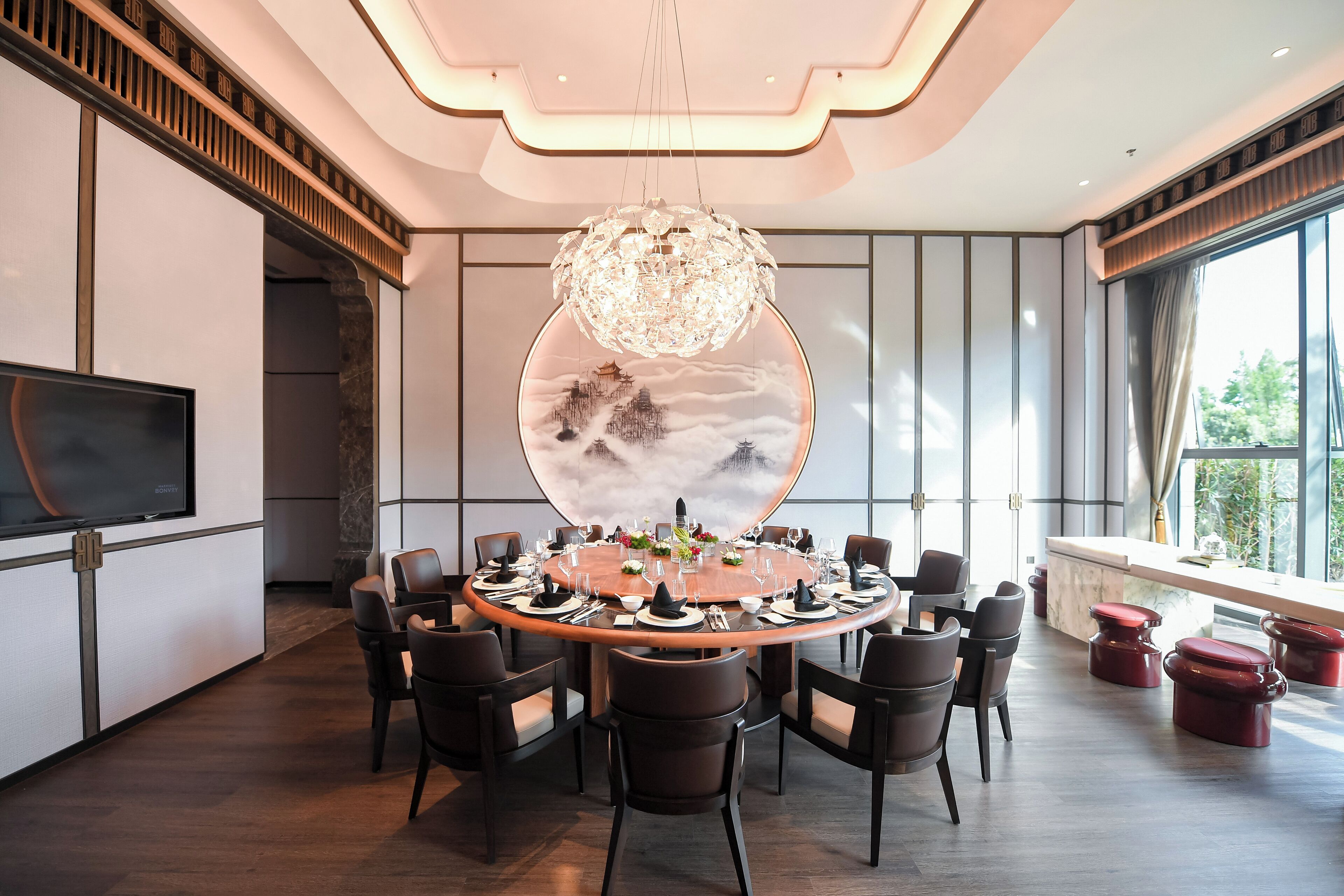 A modern dining area with a round table set for a meal, surrounded by chairs, a chandelier, and a large window with a view.
