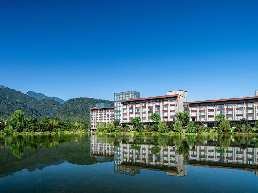 Un hotel moderno con su reflejo en el agua, rodeado de vegetación y montañas.