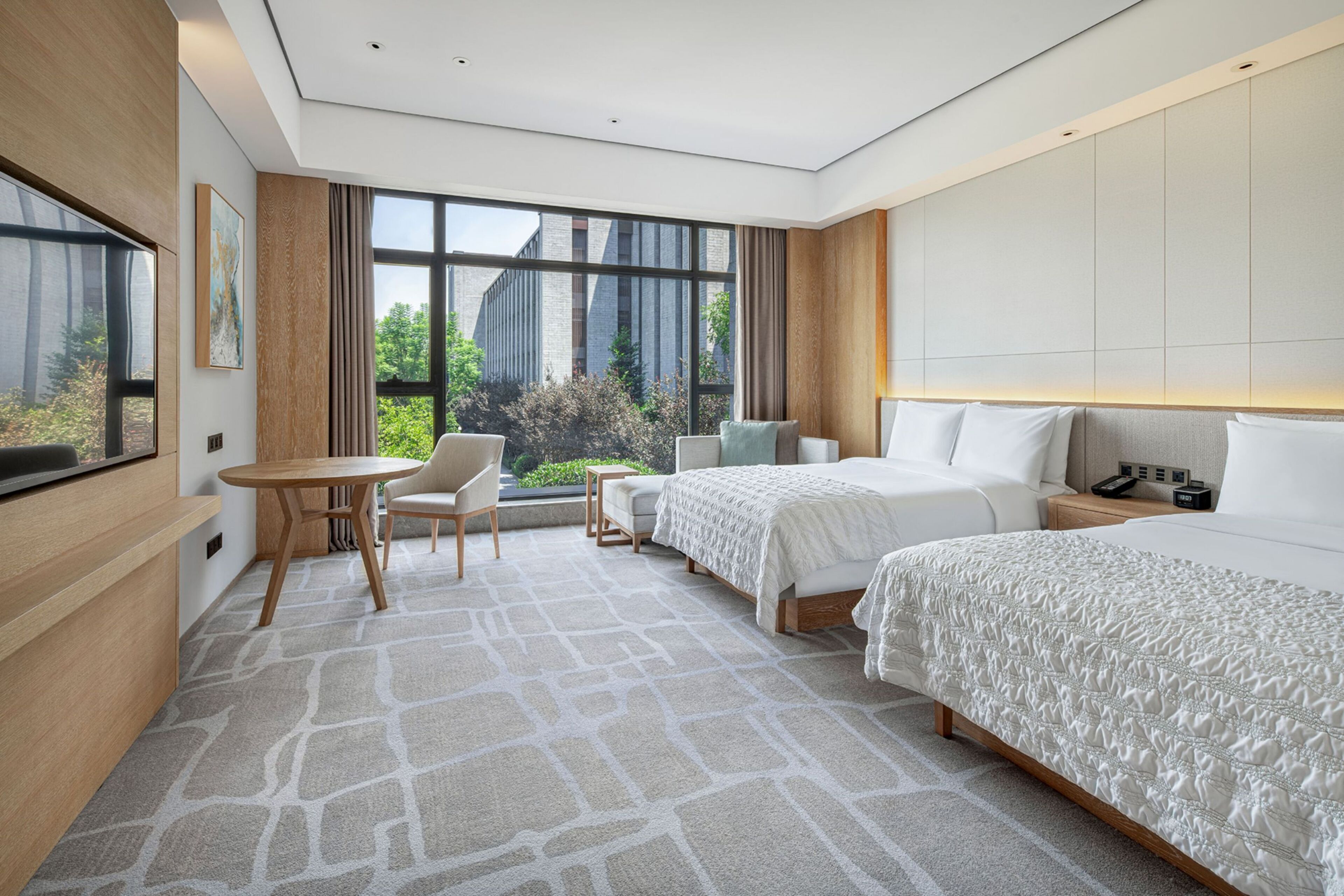 A modern hotel room with two beds, a seating area, a large window with a view of greenery, and a flat-screen TV on the left wall.