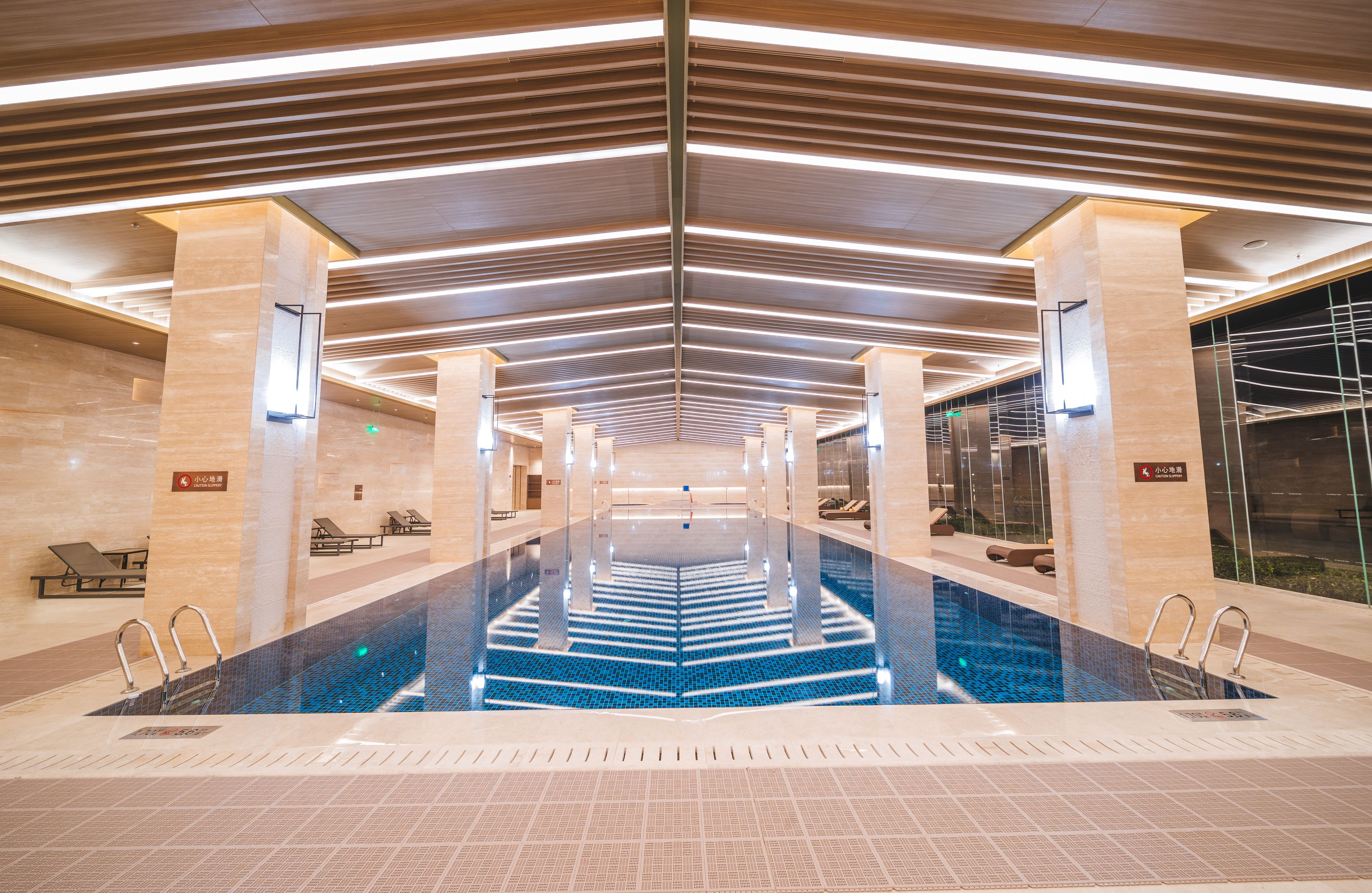 Een modern binnenzwembad met een fontein in het midden, omringd door loungegedeeltes en een hoog plafond met lineaire verlichting.