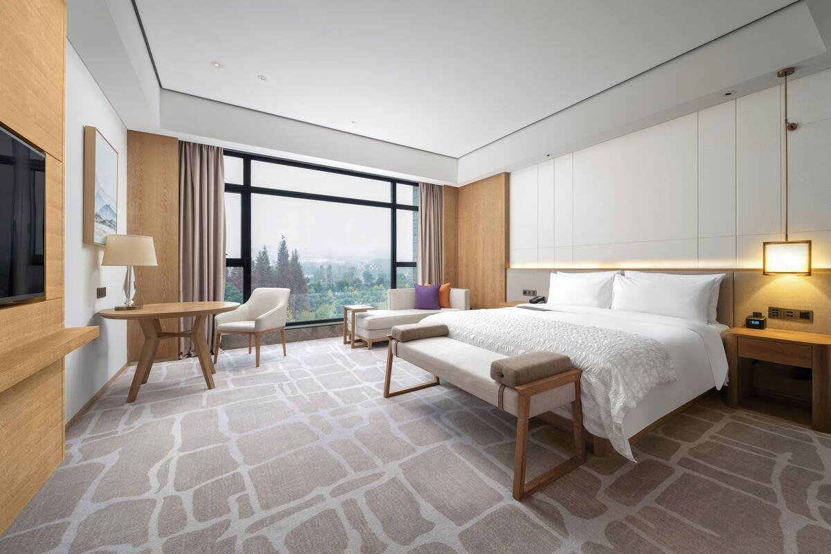 A modern hotel room with a large bed, a sitting area with a table and chairs, a television, and a view of the outdoors.
