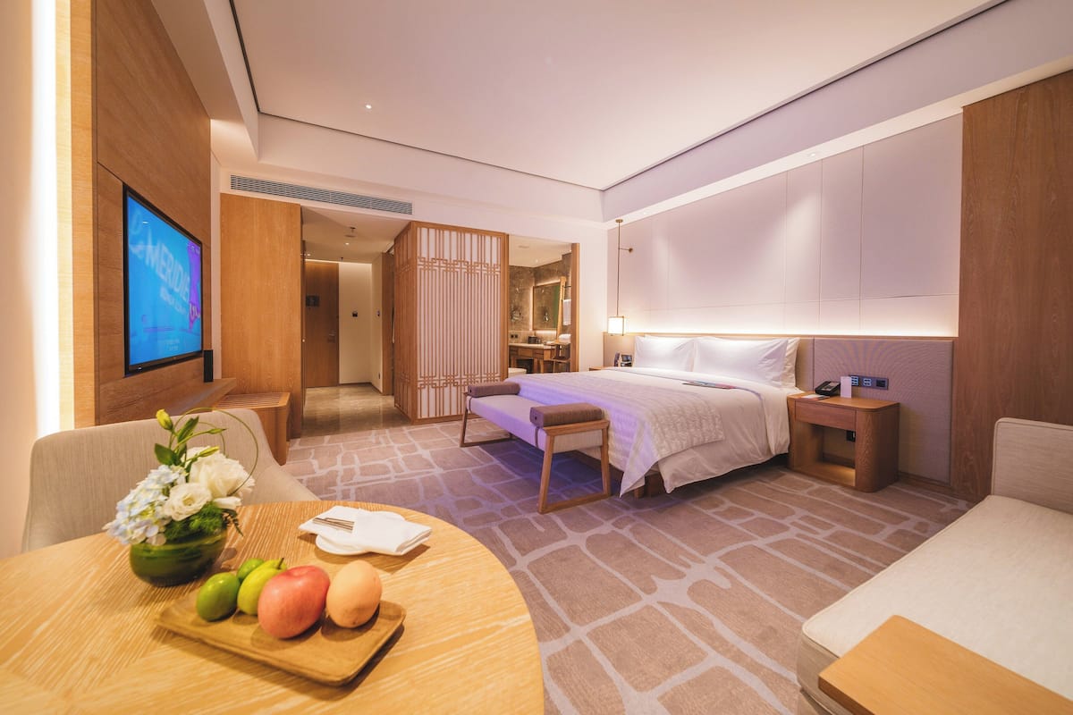 A modern hotel room with a bed, bedside table, and a TV on the wall. There is a dining area with a table and chairs, and a decorative vase with flowers.