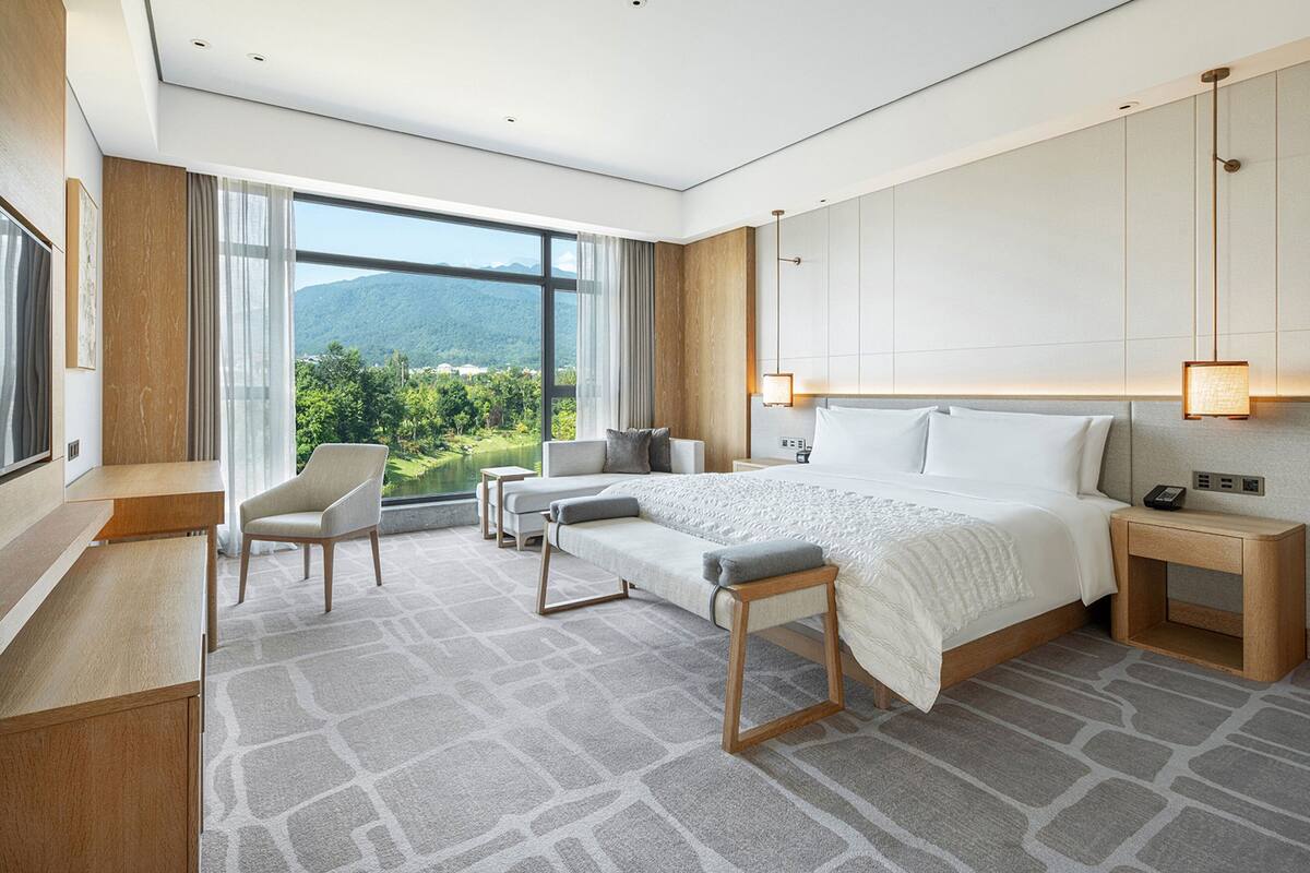 A modern hotel room with a large bed, a desk, a chair, and a view of the mountains.