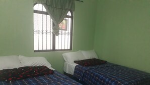 Una habitación con dos camas, cada una con colcha a cuadros azules y rojos. Se ve una ventana con reja de metal y cortina.
