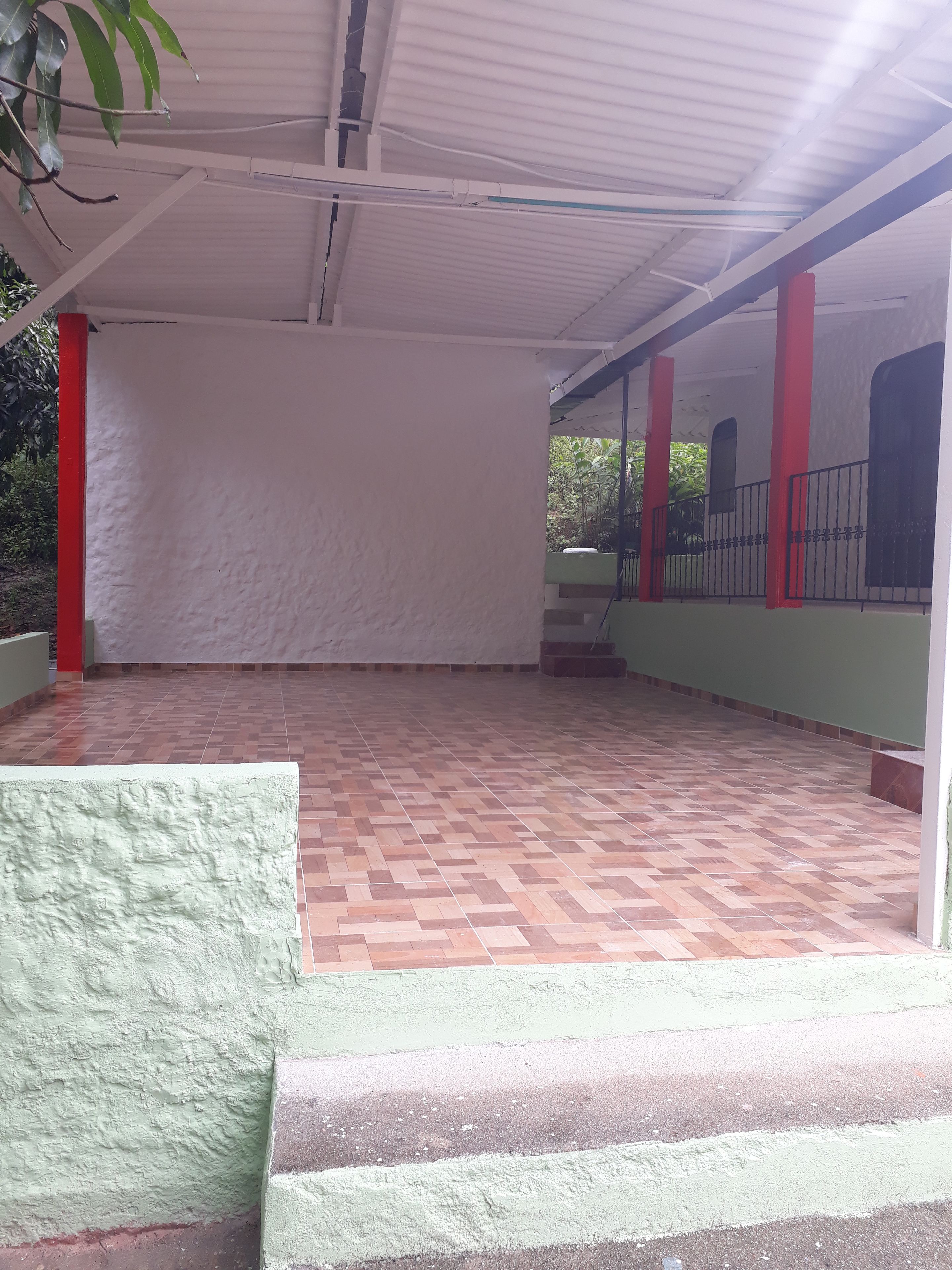 Uma área externa coberta com piso de azulejos, paredes brancas e pilares vermelhos.