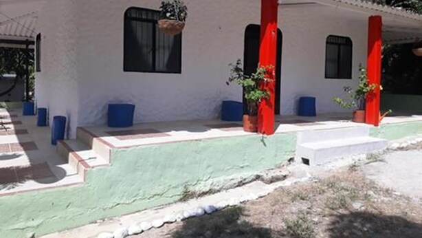 Un edificio blanco con sección inferior verde y pilares rojos. Hay escalones que conducen a un porche cubierto con cojines azules y plantas en macetas.