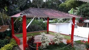 Un pequeño pabellón de techo rojo con valla de alambre, rodeado de plantas y árboles.
