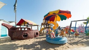 Un espace de jeux sur la plage avec une structure de jeu en forme de bateau de pirates, un parasol coloré et un petit toboggan aquatique.