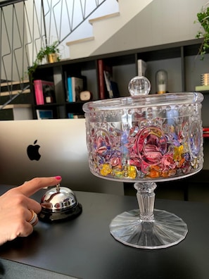 A clear glass dessert bowl with colorful candies on a table, a laptop with an Apple logo, and a hand with a bell. - Hotel Liane (Riccione)