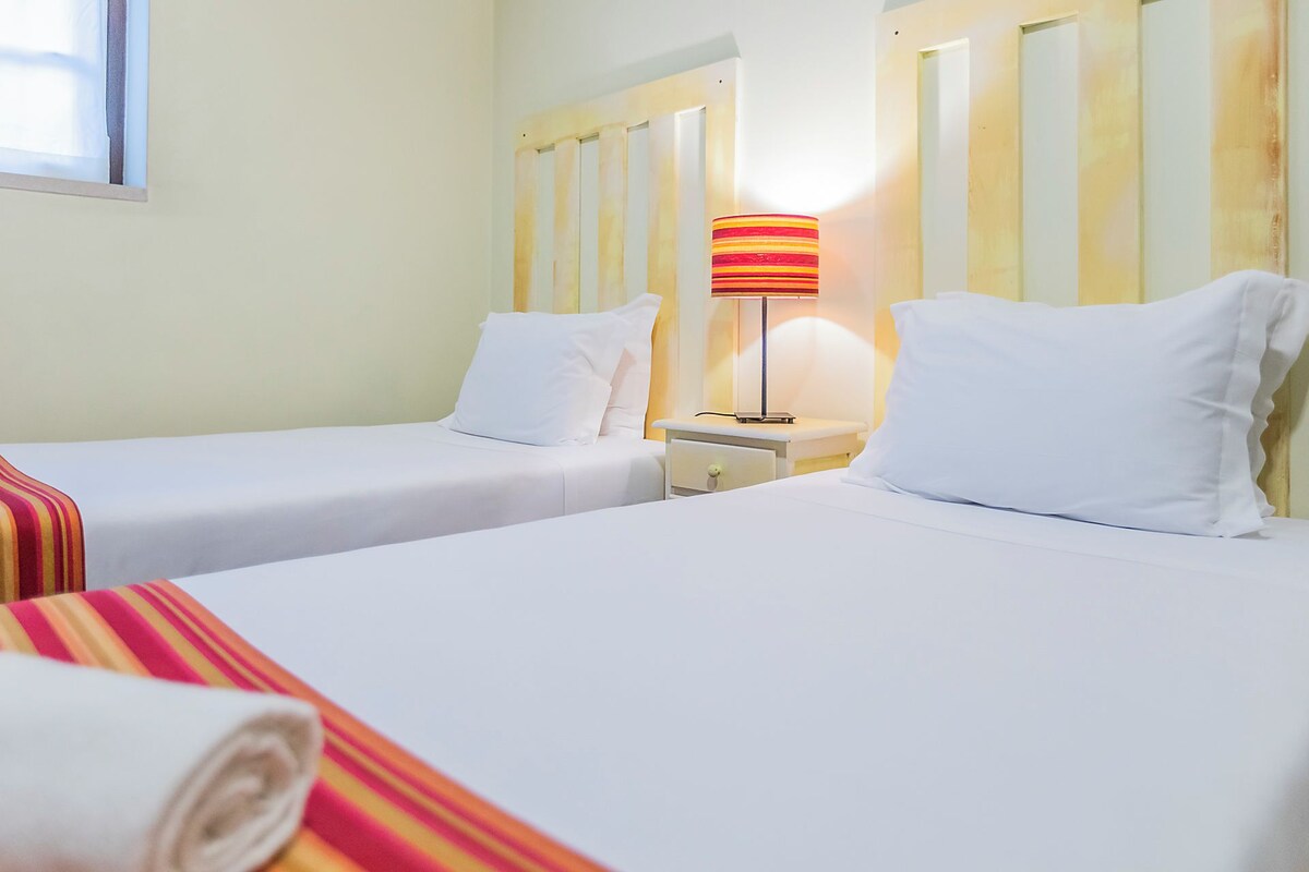 A hotel room with two single beds, a nightstand with a lamp, and a window with blinds.