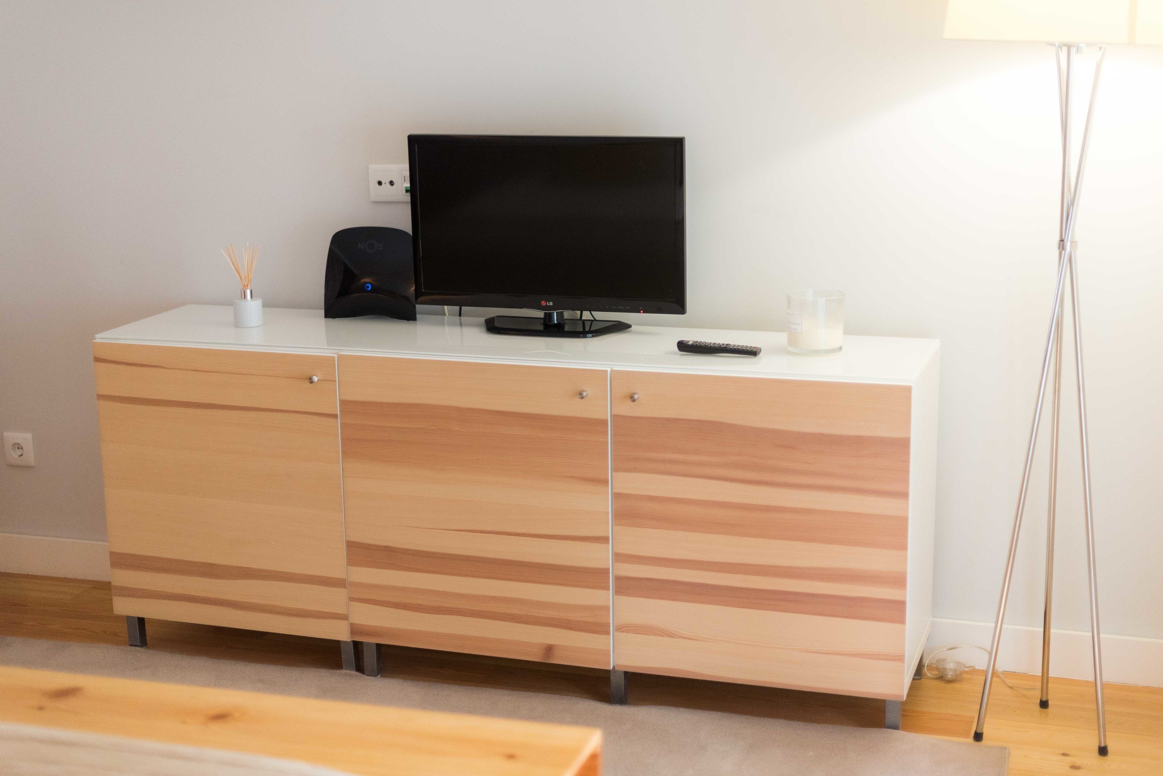 A modern living room with a flat-screen TV on a white cabinet, a remote control, a candle, and a floor lamp.