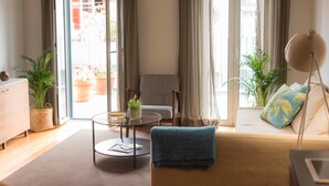 Un salon moderne avec un canapé, une table basse ronde, un fauteuil et un lampadaire. On y trouve des plantes en pot et une vue à travers une porte vitrée.