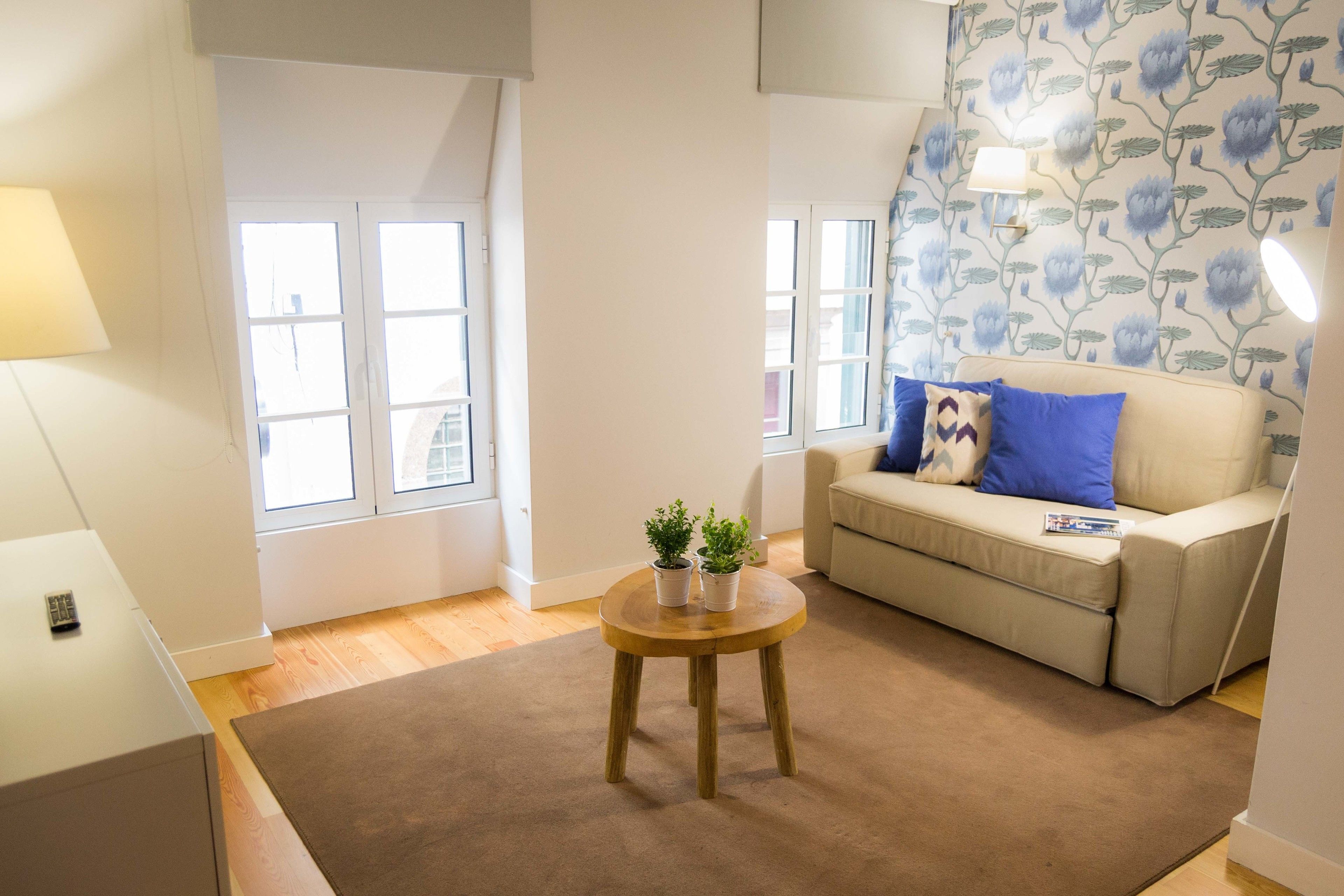 A living room with a beige sofa, two potted plants, a wooden coffee table, and a wall-mounted lamp.