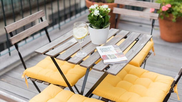 Une table en bois avec des coussins jaunes, un verre de vin blanc, une plante en pot et un magazine sur un balcon.
