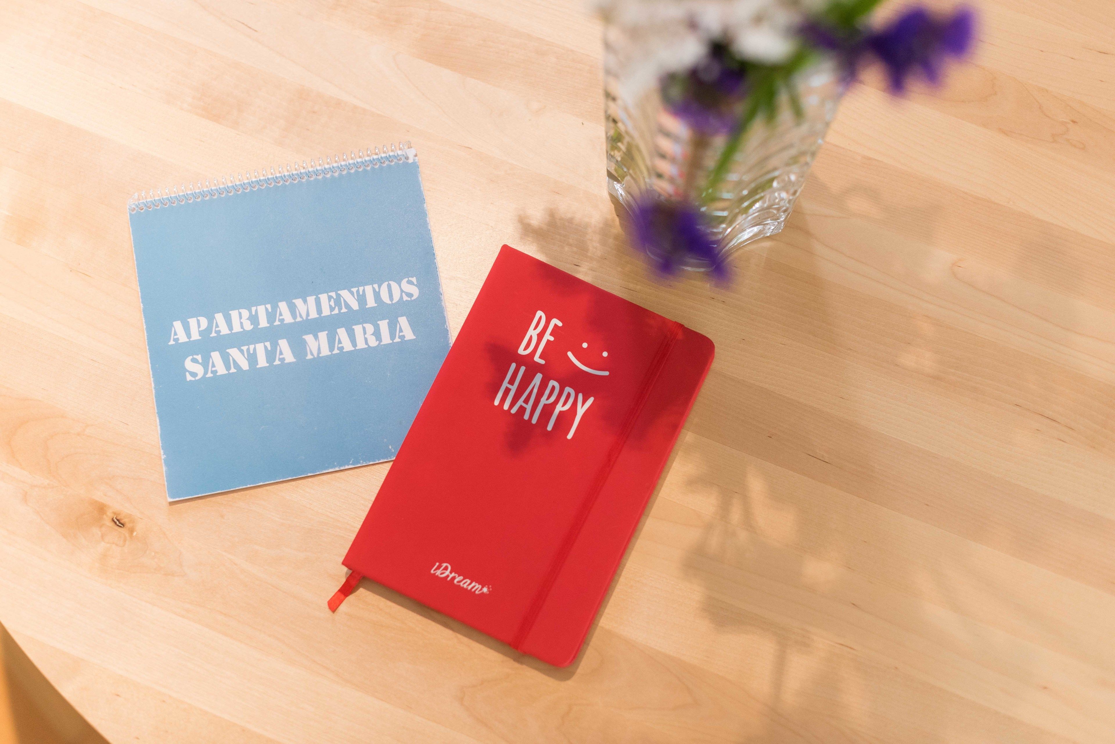 Deux cahiers sur une table en bois, l’un bleu avec l’inscription « APARTAMENTOS SANTA MARIA » et l’autre rouge avec l’inscription « SOYEZ HEUREUX ».