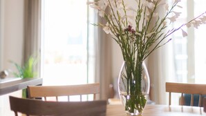 Une table à manger sur laquelle est posé un vase transparent contenant des fleurs blanches et des tiges vertes, dans une pièce dotée d’une grande fenêtre et de rideaux.