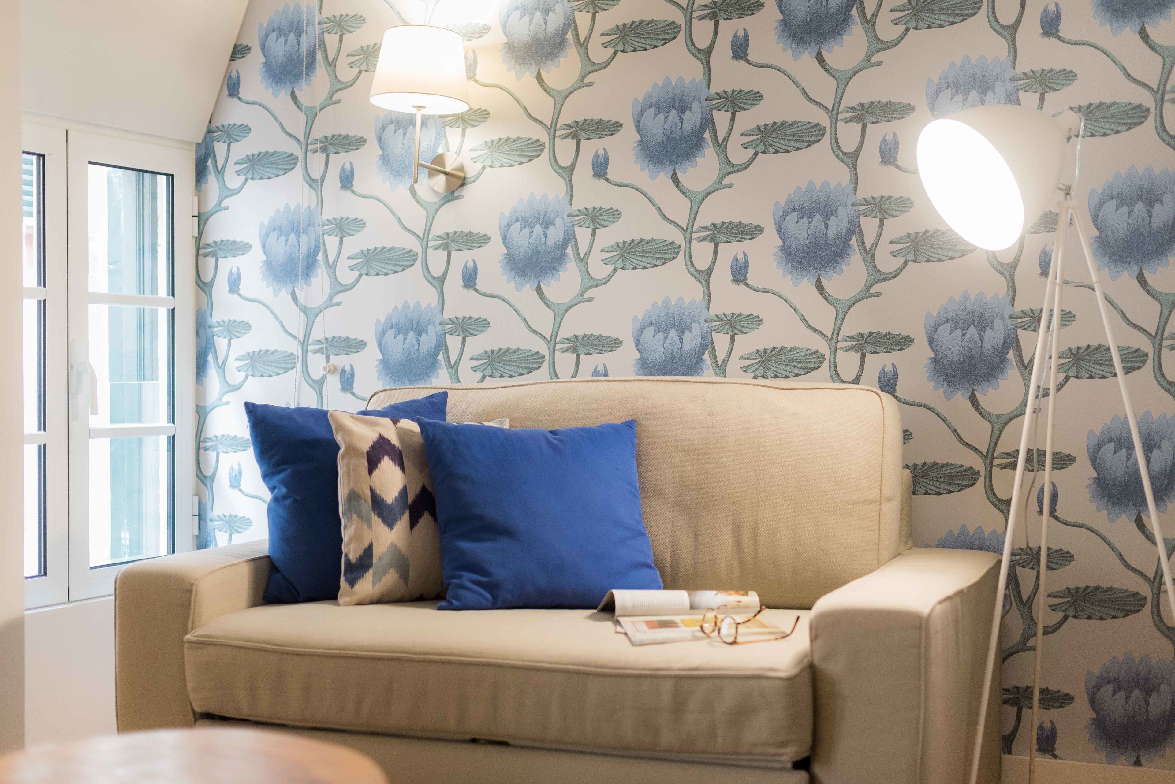 A room with a beige sofa, blue pillows, a lamp, and a wall with floral wallpaper.