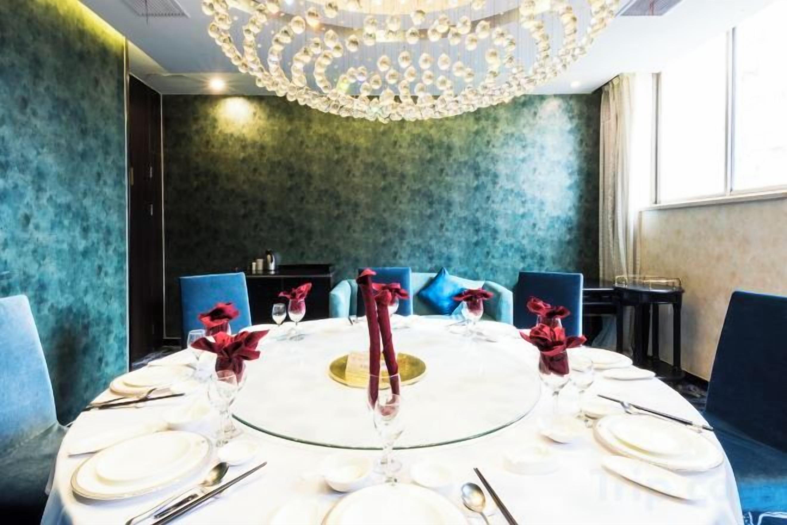 A round table set for dining with a chandelier above, blue chairs, and a green textured wall.