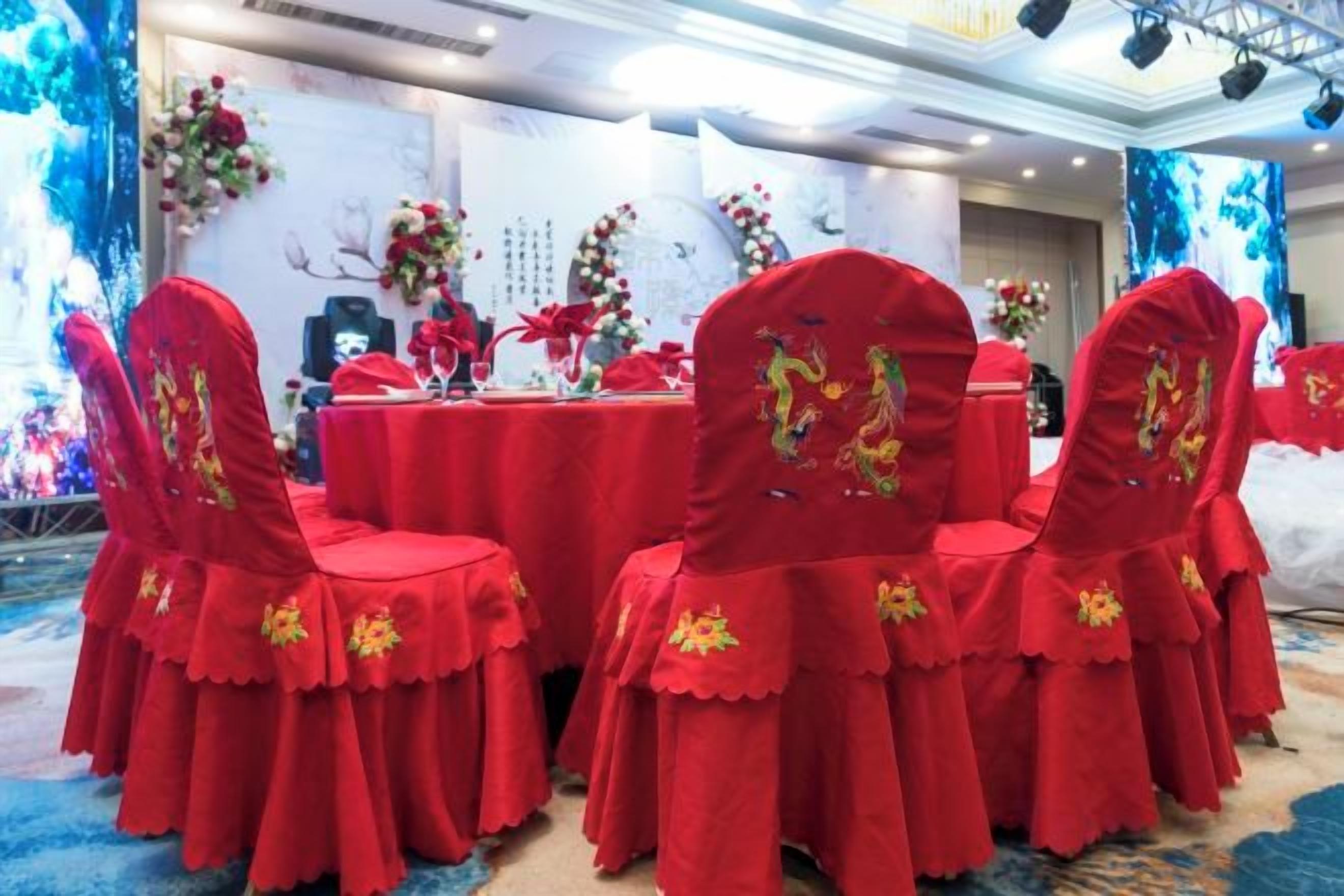 A banquet hall with round tables and chairs, decorated with floral arrangements and red tablecloths.