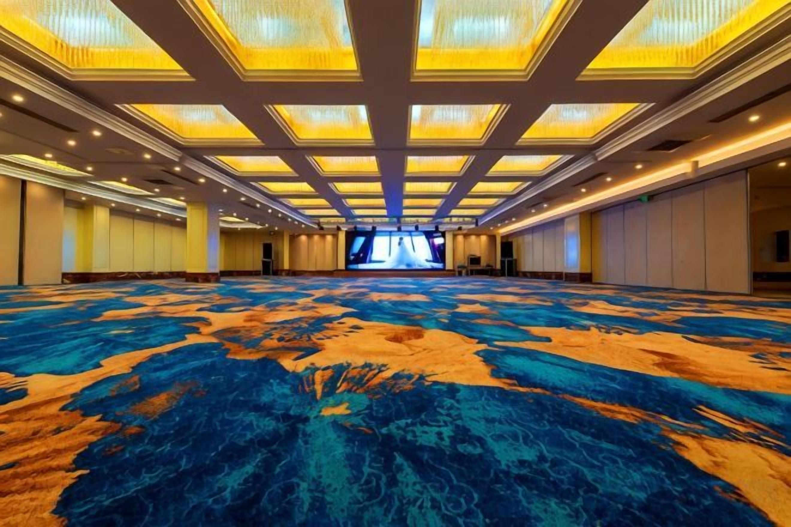 A large conference room with a patterned carpet, a high ceiling with recessed lighting, and a large screen at the front.