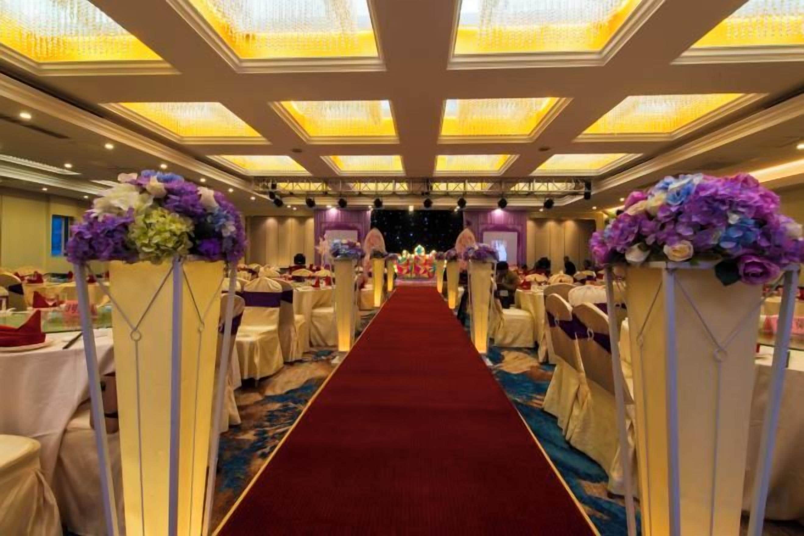 A banquet hall with a red carpet aisle, round tables, and floral centerpieces.