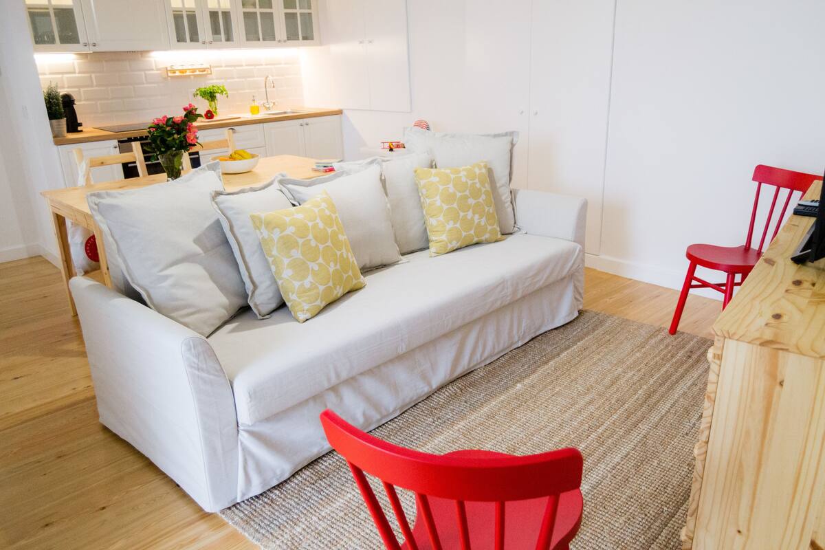 A modern living room with a white sofa, red chairs, a wooden dining table, and a kitchen in the background.