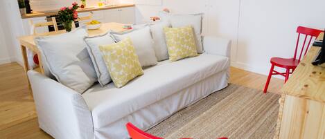 A modern living room with a white sofa, red chairs, a wooden dining table, and a kitchen in the background.