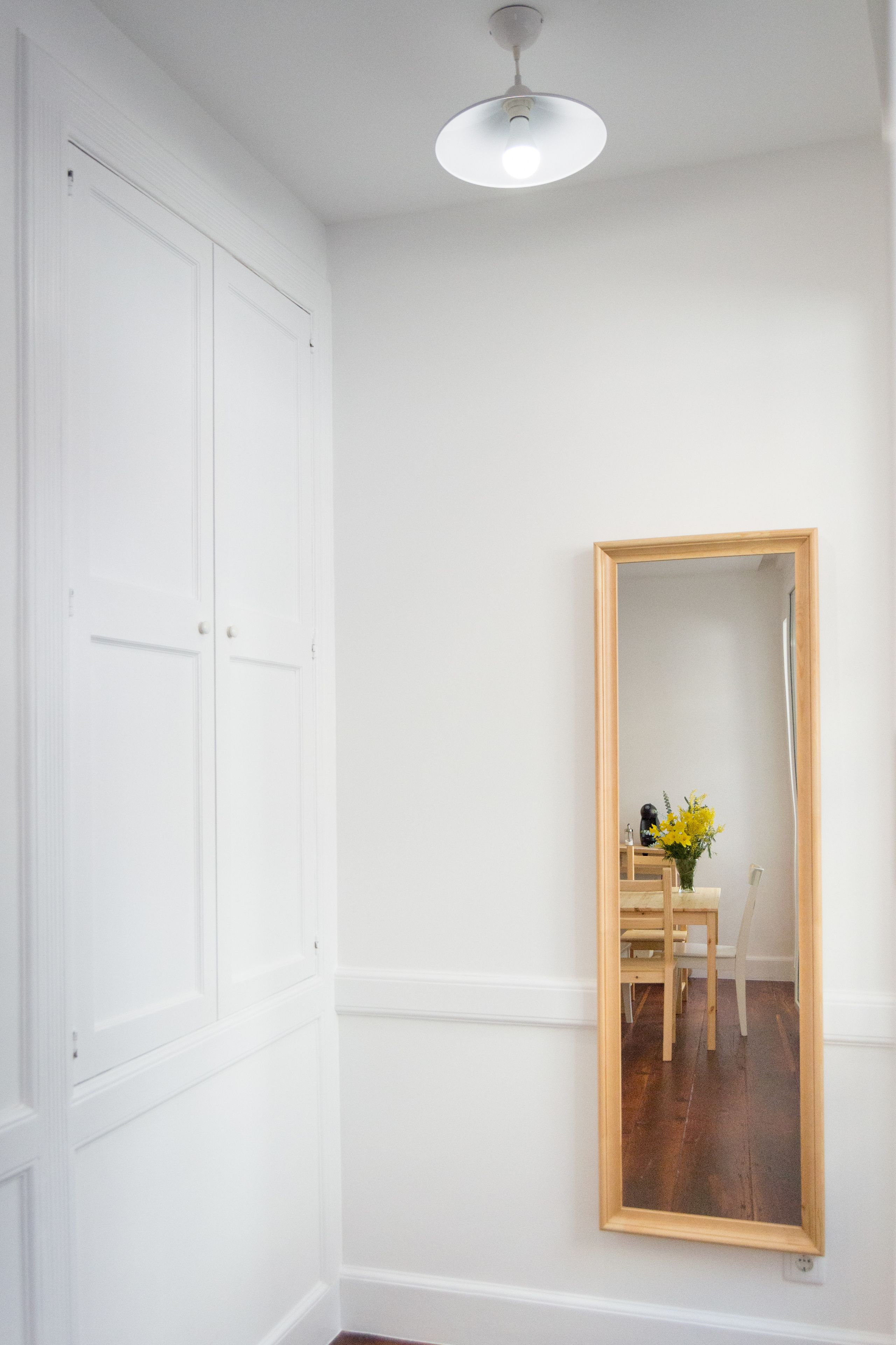 Une pièce aux murs blancs, avec un miroir à cadre en bois, un luminaire au plafond et un plancher en bois.