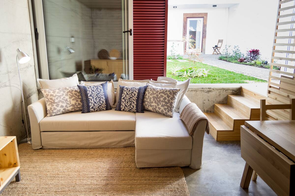 A modern living room with a beige sectional sofa, patterned cushions, a wooden coffee table, and a view of the garden through large windows.