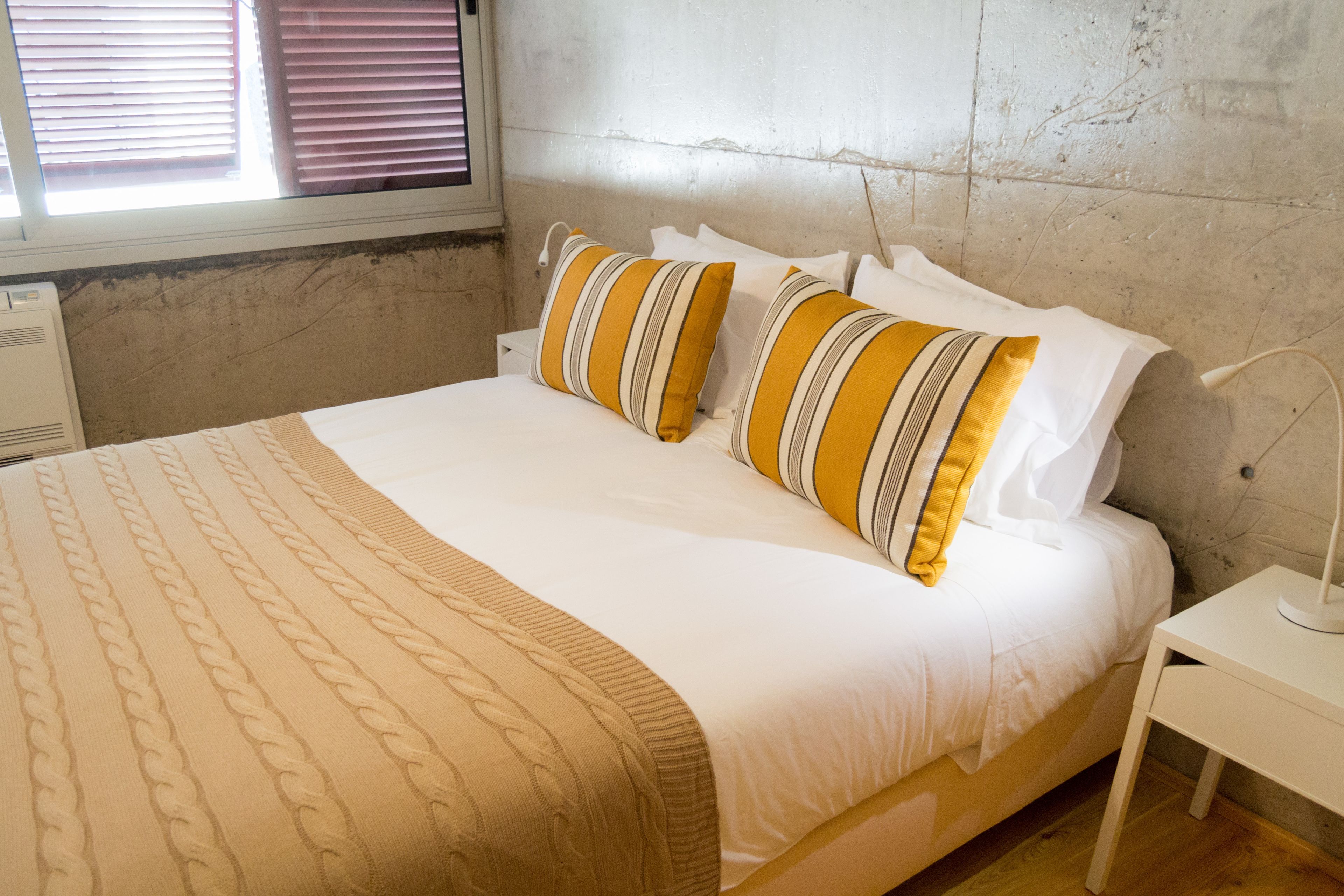 A neatly made bed with striped pillows, a white bedside table, and a window with blinds.