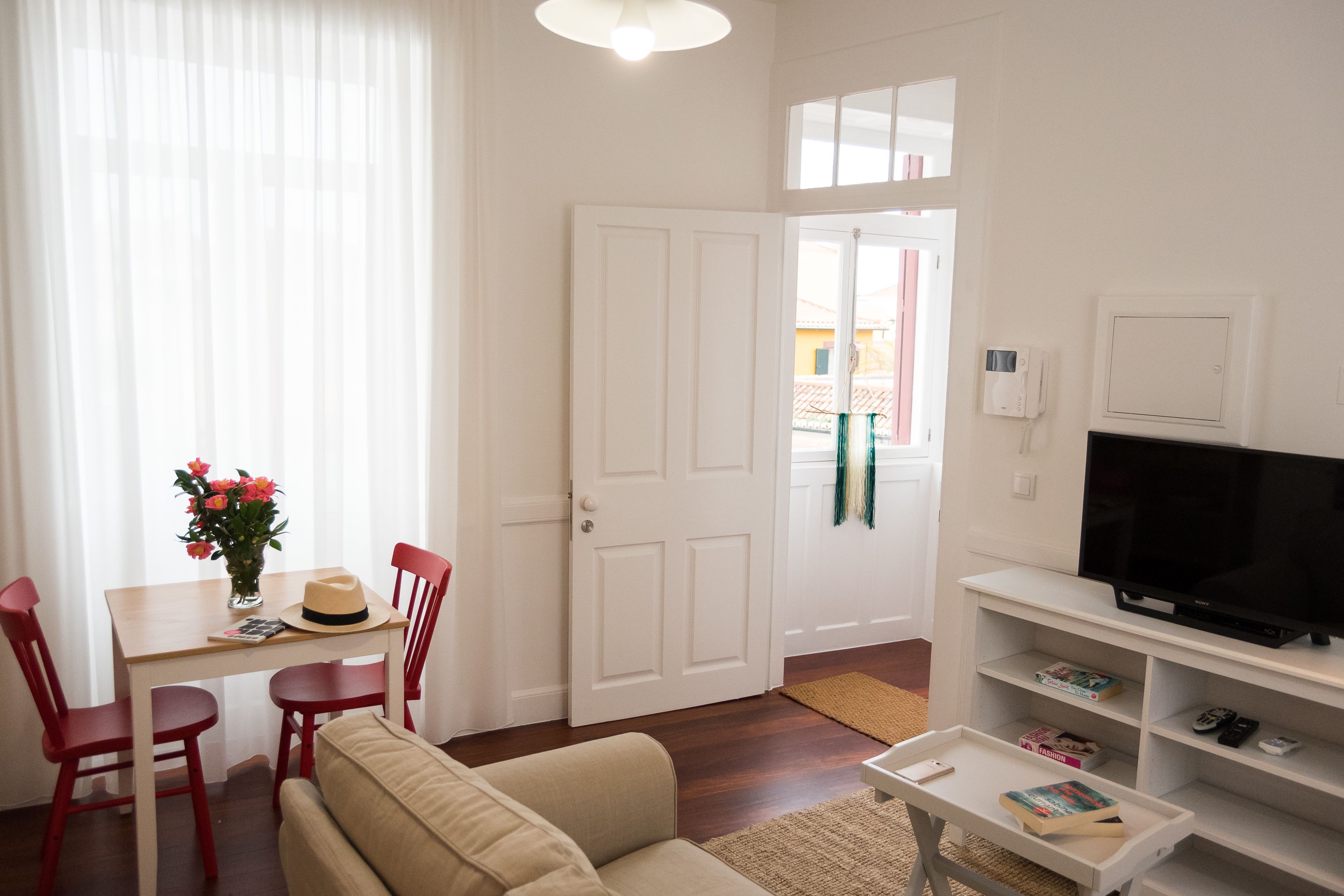 A living room with a sofa, two red chairs, a dining table with a vase of flowers, a television on a stand, and a bookshelf.