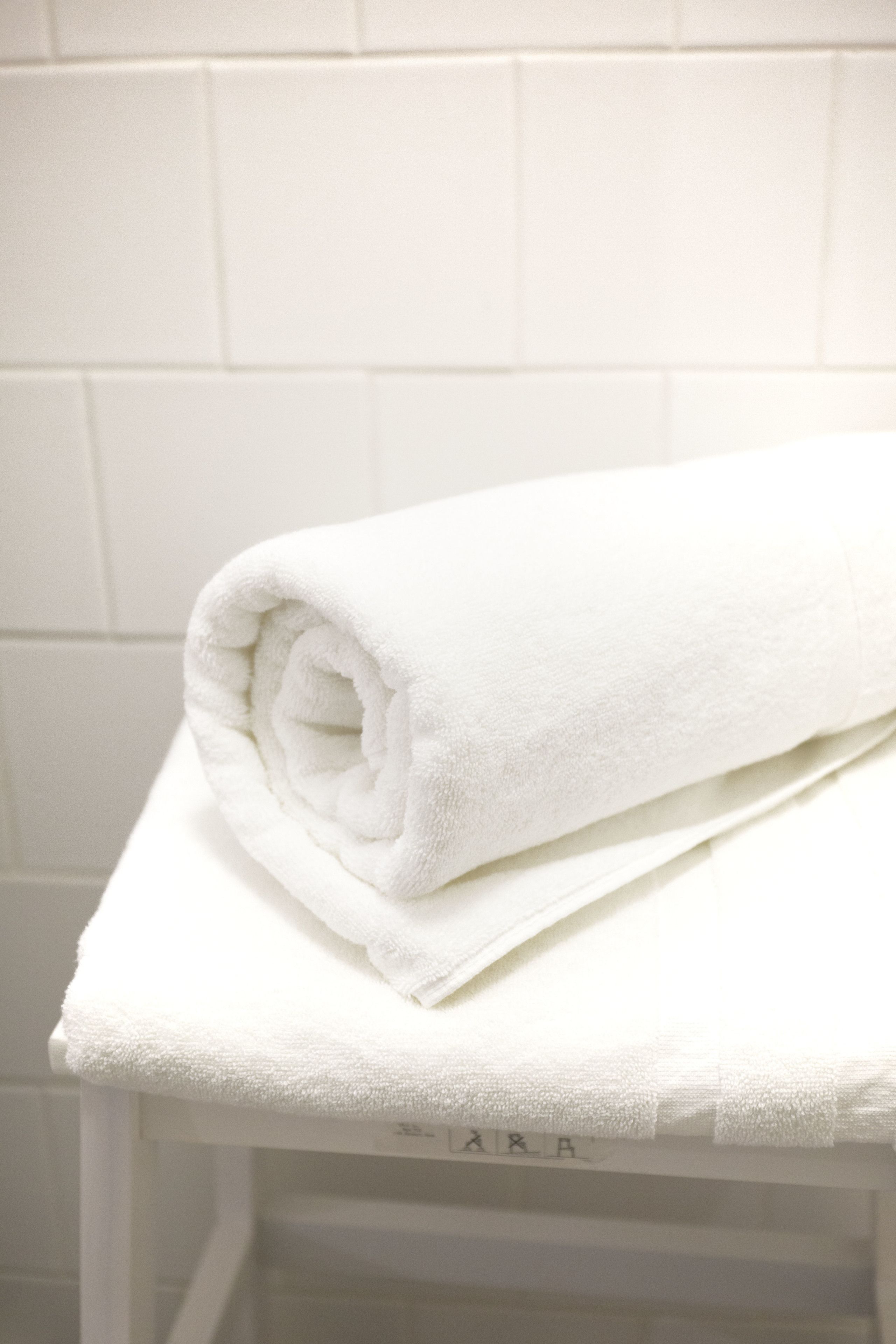 A white towel rolled up and placed on a white table in a bathroom with white tiled walls.