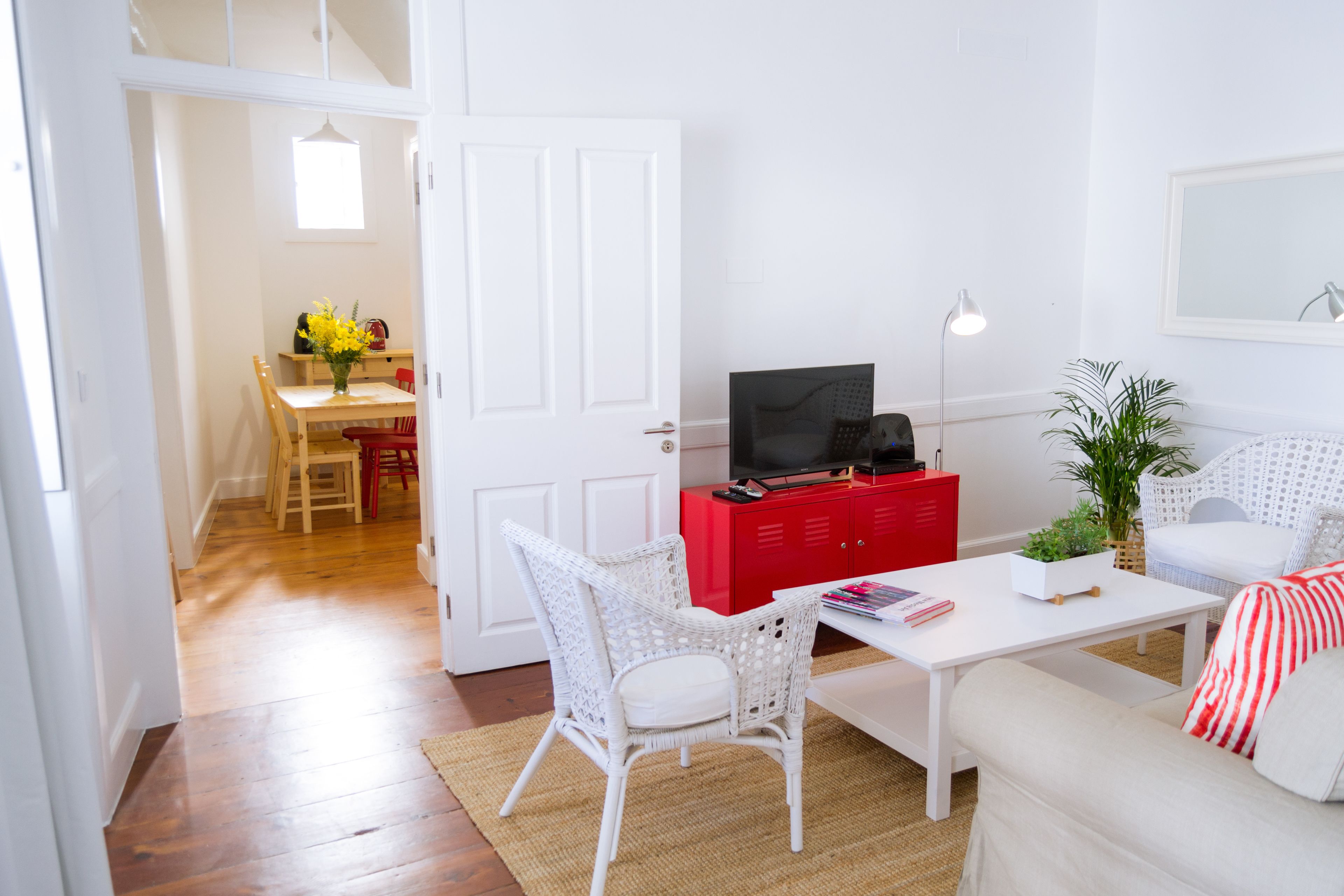 Una sala de estar con un sofá blanco, una silla de mimbre blanca, un mueble rojo con un televisor, una mesita con una planta y un acceso a otra habitación.