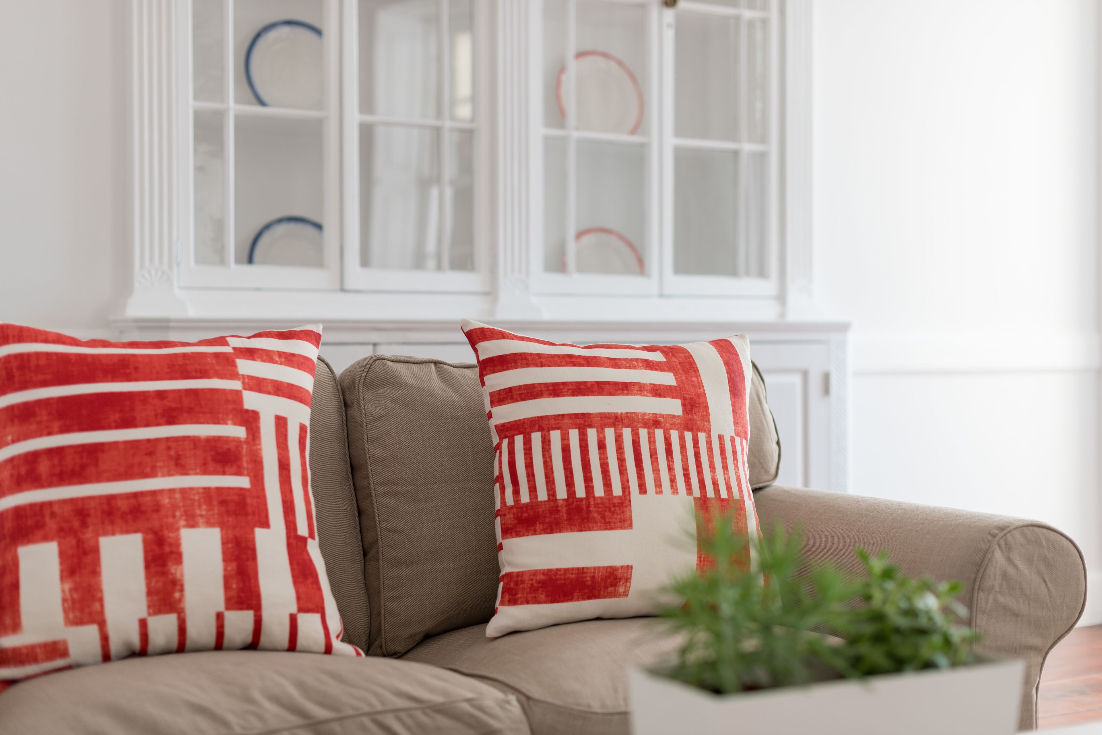 A beige sofa with red and white patterned pillows and a green plant in a white pot.