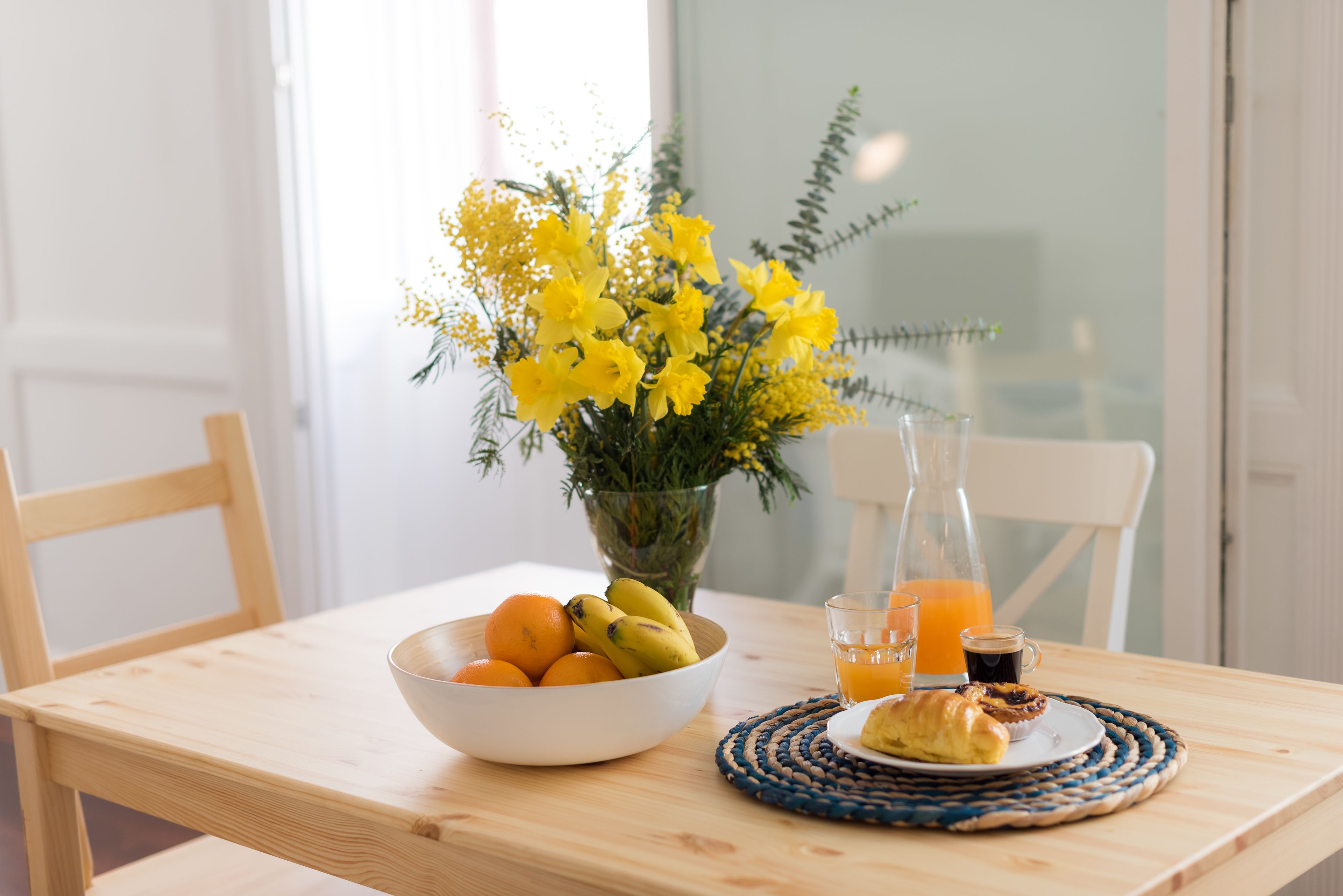 Una tavola per la colazione con una ciotola di frutta, un vaso di fiori gialli, una bottiglia di succo d'arancia e un cornetto.