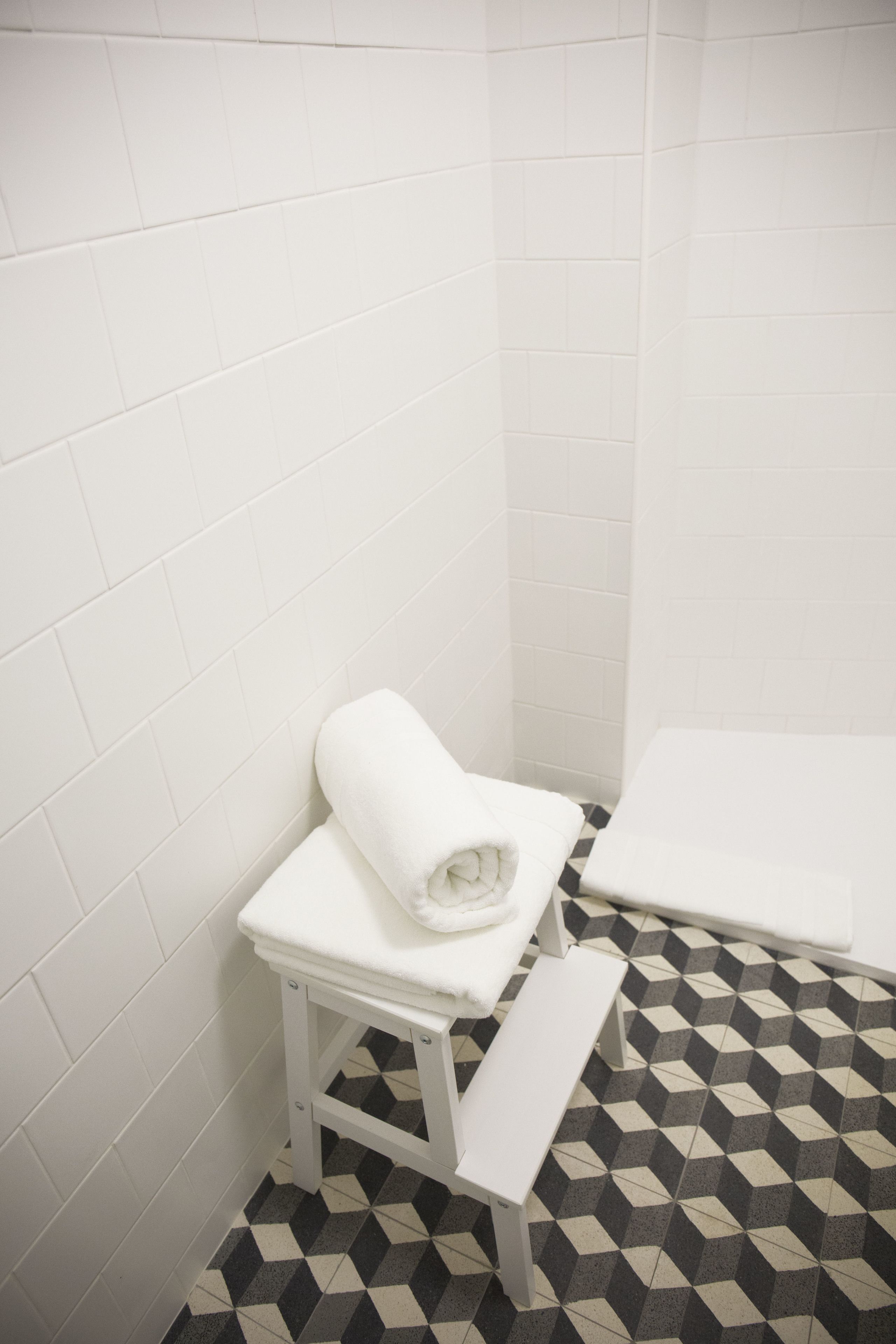 A bathroom with white tiled walls and floor, a white bench, and a rolled white towel on the bench.