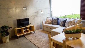 A living room with a TV on a wooden stand, a white sofa with patterned cushions, a wooden table with a plant, and a large window with a view of greenery. - Casa Do Mercado (Funchal)