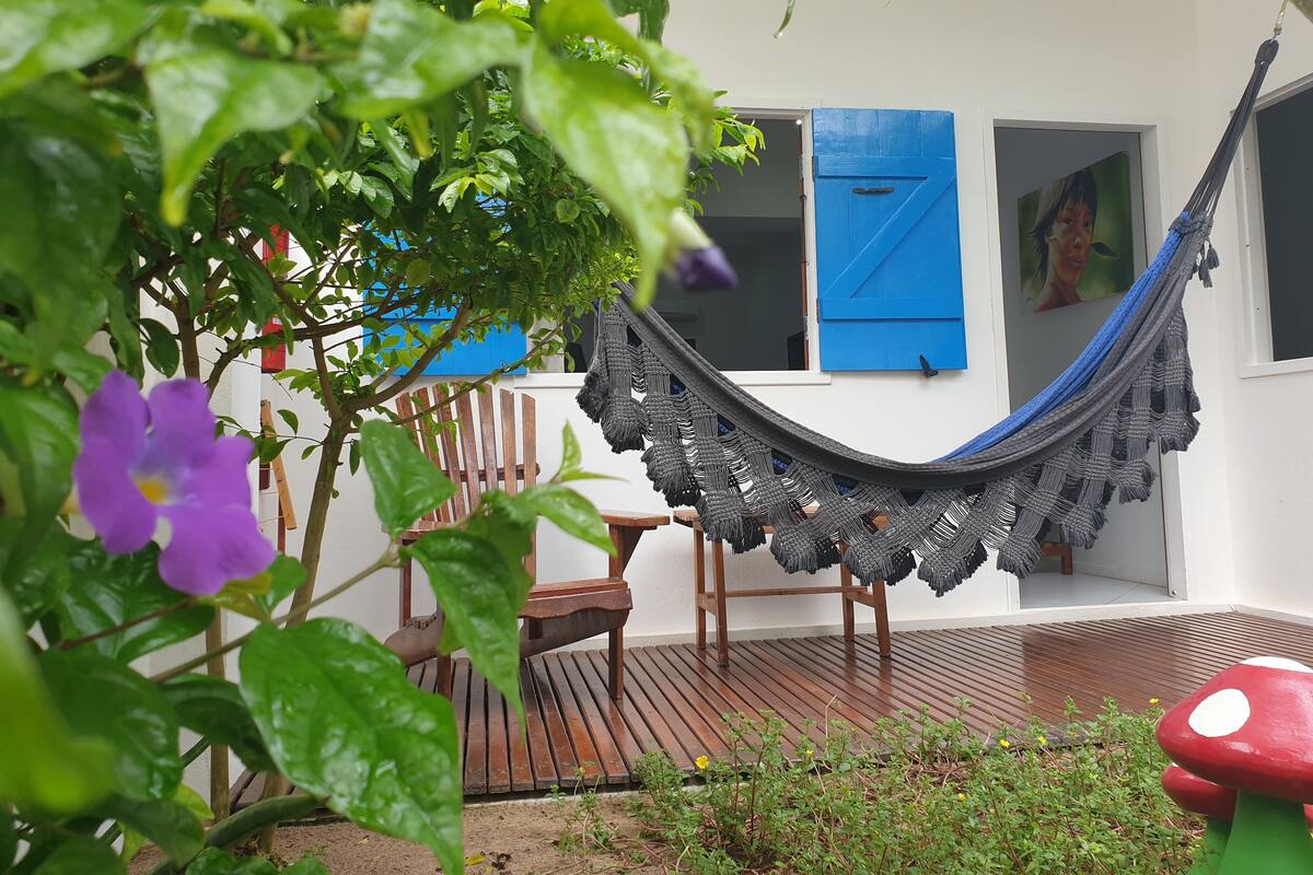 A hammock hanging between two windows with blue shutters, a wooden deck, and a garden area with plants and flowers.