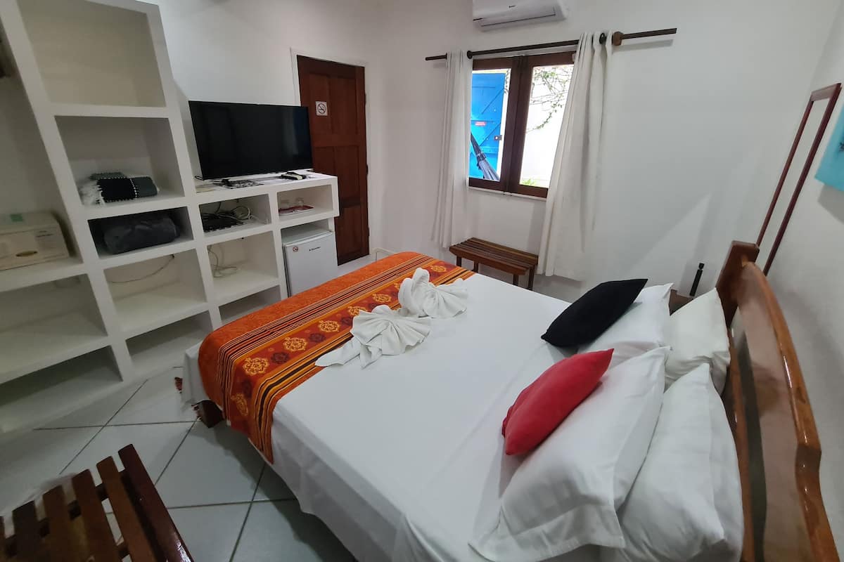 A hotel room with a bed, a TV on a stand, a wardrobe, a window with curtains, and an air conditioning unit.