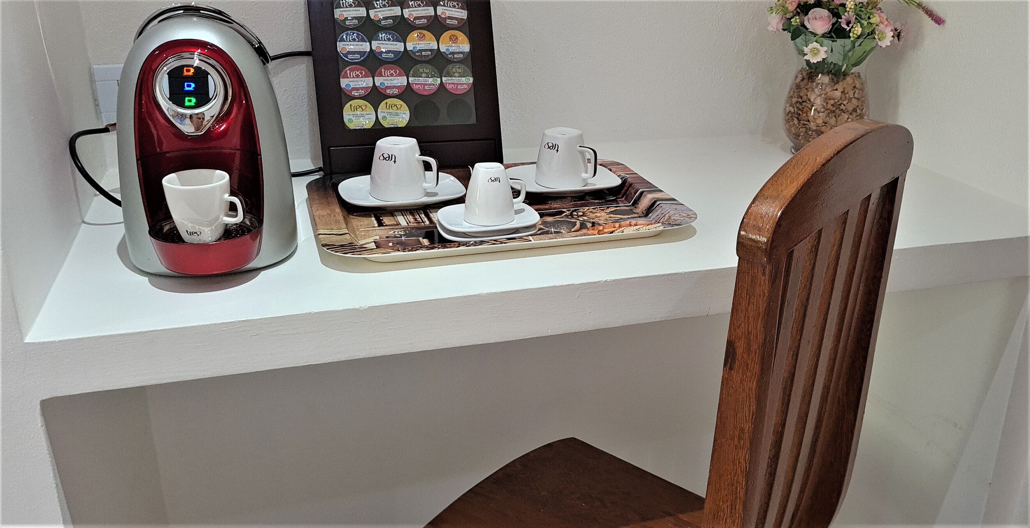 A coffee machine with a digital display, a tray with cups and saucers, and a small vase with flowers on a white table.