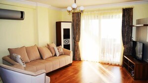 Un soggiorno con un divano color beige, una poltrona bianca, un mobile TV in legno e una grande finestra con tende.