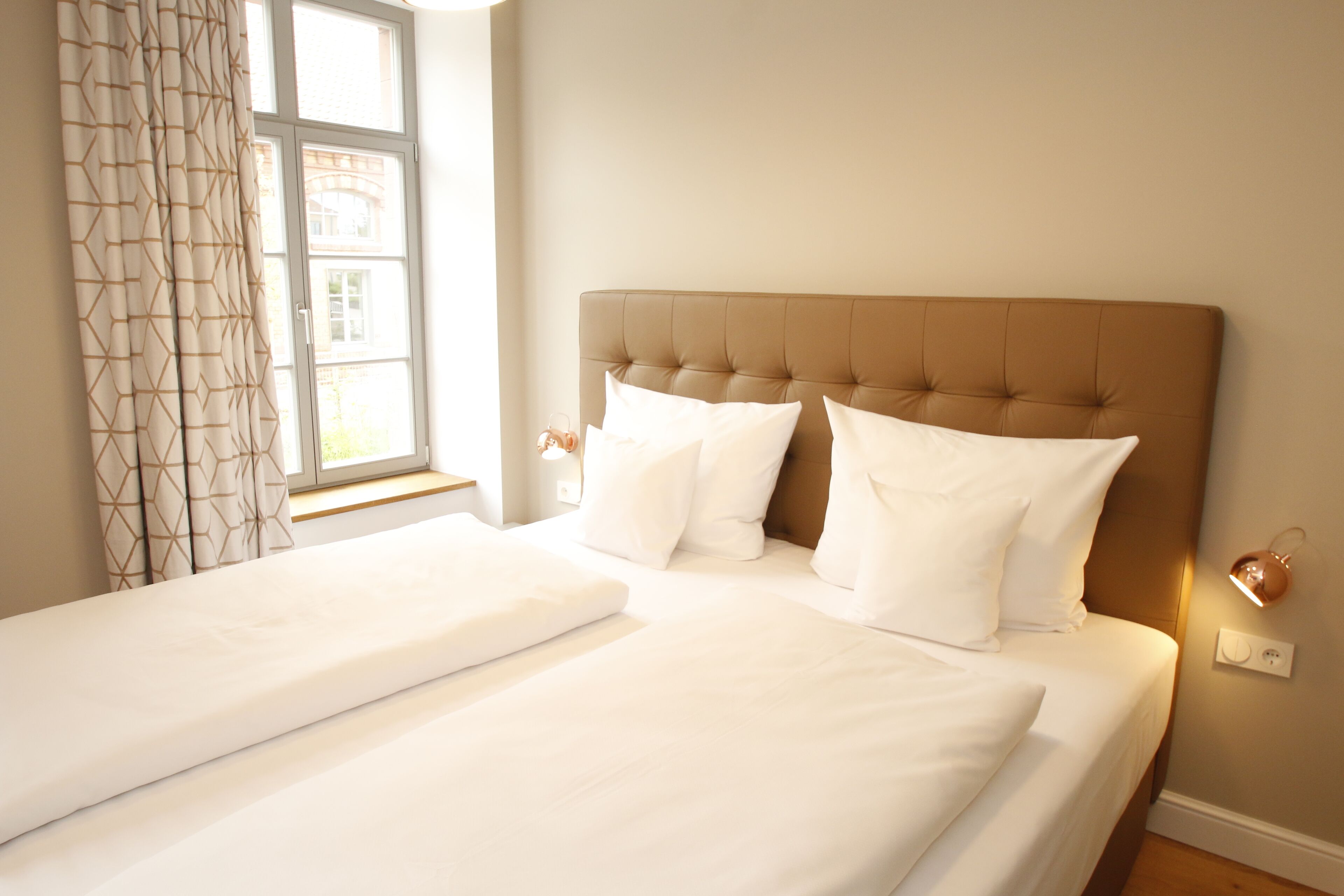 Ein Hotelzimmer mit einem großen Bett, zwei Kissen, einem Fenster mit Vorhängen und einer Nachttischlampe.